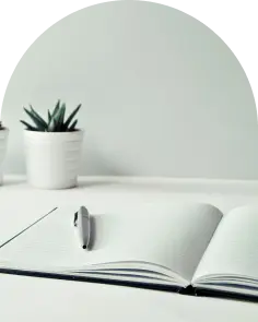 Photo d'un livre ouvert avec un stylo posé sur une page et une plante en arrière plan